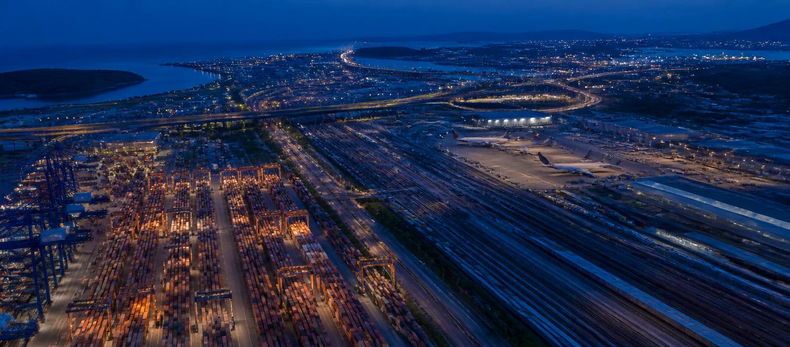 Aerial view at dusk of a multimodal hub where a container yard, rail sidings and an aircraft apron meet