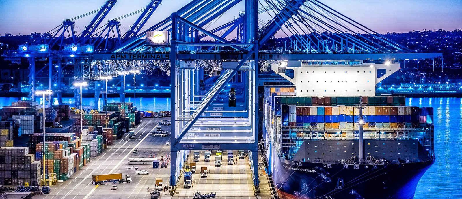Container vessel alongside a floodlit terminal at dusk while gantry cranes work the stacks